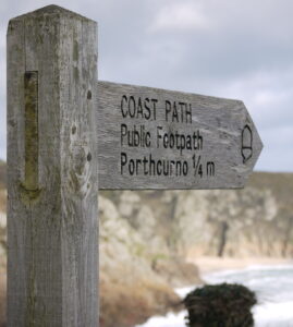 A wooden Coast Path footpath sign to Porthcurno Cornwall. Part of the South West Coast Path walk. view of the cliffs of Porthcurno in the background.